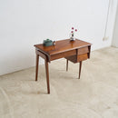 A mid-century modern wooden writing desk with a warm brown finish, featuring a single drawer, tapered legs, and small decorative objects on top, set against a plain white wall and light concrete floor.