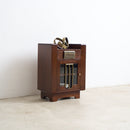 A converted Art Deco radio cabinet with a glass-front lower door, brass handle, and a small gold-toned object on top, set against a plain white wall and light concrete floor.