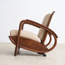 An Art Deco armchair with handcarved teak armrests, cream-colored upholstered seat and back cushions, and a dark teak wood base, photographed against a plain white wall and light concrete floor.