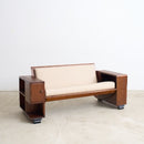 An Art Deco wooden loveseat with cream-colored cushions, featuring integrated side tables on both ends, set against a plain light gray wall and concrete floor.