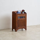 A small vintage-style wooden cabinet with a curved top, single door, and decorative window panels, shown in a minimalist room with a white wall and light concrete floor.