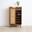 A mid-century modern wooden shoe cabinet with a woven cane door panel, open shelves, and tapered legs, styled with a small vase of flowers and shoes on top, set against a plain white wall and concrete floor.