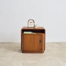 An Art Deco wooden nightstand with a warm brown finish, featuring a single cabinet door, open top shelf, and a small round clock and mug on top, set against a plain white wall and concrete floor.