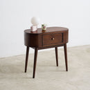 A mid-century modern wooden side table with a single drawer and tapered legs, styled with a white globe lamp and a small cup on top, set against a minimalist white wall and concrete floor.