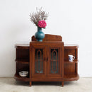 A vintage wooden sideboard cabinet with curved sides and glass-paneled doors, styled with a blue vase of pink flowers on top and decorative items on the shelves, set against a plain white wall.