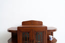 An Art Deco wooden cabinet with a warm reddish-brown finish, featuring glass-paneled doors and curved side shelves, shown against a plain white wall with a small blue and white mug on the lower shelf.