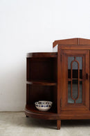 A dark brown wooden corner cabinet with a glass-paneled door and open shelves, shown against a plain white wall and light concrete floor.