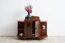 A vintage dark brown wooden sideboard cabinet with glass doors, styled with a blue vase of pink flowers on top, set against a plain white wall and light concrete floor.
