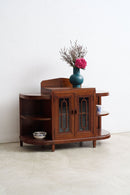 An Art Deco wooden sideboard cabinet with curved shelves and glass-paneled doors, styled with a teal vase of pink flowers on top, set against a plain white wall and light concrete floor.