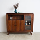 An Art Deco wooden sideboard cabinet with a warm brown finish, featuring two lower doors, an open shelf, and a glass-front cabinet door, styled with a blue vase on top against a white wall.