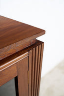 Close-up detail of An Art Deco wooden cabinet corner, showcasing a rich walnut-like brown wood grain, a fluted side panel, and a brass hinge against a white wall.