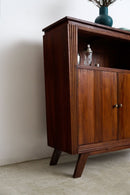 An Art Deco sideboard with a warm reddish-brown finish, featuring a top shelf, two cabinet doors with decorative grooved panels, and angled legs, shown in a minimalist room setting.