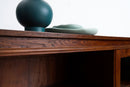 A close-up of a dark brown wooden cabinet or table with a dark teal spherical vase and a light green dish on top, set against a plain white background.