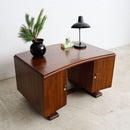 An Art Deco wooden desk with a curved top, featuring two cabinet doors on the right, a center opening, and a dark wood grain finish. The desk is styled with a black lamp, a small green box, and a potted pine branch, set against a plain white wall and light concrete floor.
