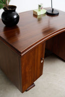 An Art Deco teak writing desk with a rich reddish-brown finish, featuring a cabinet door with a small silver knob, shown in a home office setting with a black vase, books, and a lamp on top.
