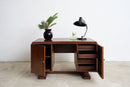 An Art Deco wooden writing desk with open doors showing drawers and shelving, styled with a black lamp, a small plant in a black pot, and a cup on top, set against a plain white wall and light concrete floor.