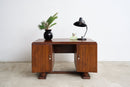 An Art Deco  wooden writing desk with a warm brown finish, styled with a black vase of green leaves and a black desk lamp on top, set against a plain white wall and light concrete floor.