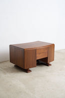 An Art Deco wooden nightstand with a warm brown finish, featuring a single drawer and open shelf, set against a plain white wall and light concrete floor.