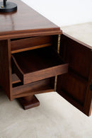 A close-up of a dark brown wooden cabinet with one drawer pulled open, showing its interior shelves, set against a light beige floor and white wall.