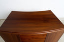 A close-up of a curved wooden cabinet or desk top made of rich reddish-brown wood grain, set against a plain white wall and light floor.