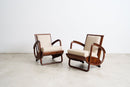 Two Art Deco Streamline Moderne wooden armchairs with cream upholstered seats and backs, shown on a light concrete floor against a white wall.