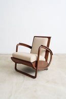 An Art Deco Streamline Moderne armchair with curved dark brown wood frames and cream-colored upholstered seat and back cushions, set against a plain light gray wall and concrete floor.