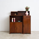 An Art Deco Sideboard in teak with a warm brown finish, featuring open shelves, a single drawer, and two lower doors. It is styled with decorative items including a pink vase with flowers, set against a plain white wall and light concrete floor.