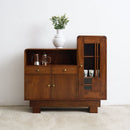 An Art Deco modern wooden sideboard cabinet with a warm brown finish, featuring a glass-front cabinet on the right, a central drawer, and an open shelf on the left. A white ceramic vase with green foliage sits on top.