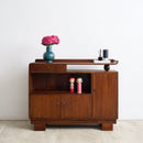 An Art Deco wooden sideboard cabinet with a warm brown finish, styled with a blue vase of pink flowers and small decorative items on top, set against a plain white wall and light concrete floor.