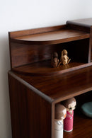 A close-up of a dark brown wooden cabinet or shelf unit with curved open compartments, holding small decorative figurines, against a plain light wall.