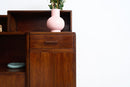 An Art Deco Sideboard with a single drawer and open shelving, styled with a pink ceramic vase containing green stems against a plain white wall.