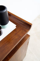 Close-up of a dark brown wooden sideboard or cabinet corner with a curved front edge, showing a white tray and black ceramic vases on top against a light neutral background.