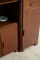 A close-up of an Art Deco Sideboard with a warm brown finish, featuring a single drawer, a lower cabinet door, and a side shelf with a small light blue dish, set against a light beige stone floor.