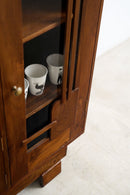 An Art Deco wooden cabinet with a glass door, showing white ceramic mugs on the shelf, set against a light-colored floor and wall.