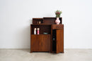 An Art Deco Sideboard with a warm brown finish, featuring open shelves, a drawer, and a lower cabinet door. It is styled with a pink vase of flowers and small decorative items on top, set against a plain white wall and concrete floor.