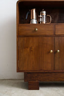 An Art Deco wooden sideboard cabinet with a warm brown finish, brass knobs, and open shelf displaying copper and glass coffee makers, set against a plain light wall and concrete floor.