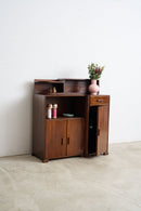 An Art Deco Sideboard cabinet with a warm brown finish, featuring open shelves, a single drawer, and a lower cabinet door, styled with decorative items and a pink vase against a white wall and concrete floor.