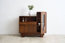 An Art Deco wooden sideboard cabinet with a warm brown finish, featuring a glass-front cabinet on the right, open shelving, and two drawers on the left. A white ceramic vase with green leaves sits on top.