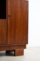 A close-up of an Art Deco sideboard 
with a warm reddish-brown finish, featuring brass hinges, a round brass knob, and a block-style leg, set against a plain white wall and concrete floor.