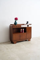 An Art Deco sideboard cabinet with a warm brown finish, featuring a top drawer, open shelf, and two lower cabinet doors. It is styled with a blue vase of pink flowers and a small desk lamp on top, set against a plain white wall and concrete floor.