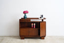 An Art Deco sideboard cabinet with a warm brown finish, featuring an open shelf with a blue vase of pink flowers, two small doll figures, and a tray on top, set against a plain white wall and light concrete floor.