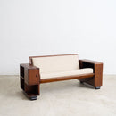 An Art Deco wooden loveseat with cream-colored cushions, featuring a built-in side table on the left, set against a plain white wall and concrete floor.