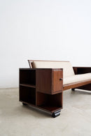 An Art Deco sofa with a dark brown wood frame, beige upholstered seat and back cushions photographed in a minimalist studio setting with a concrete floor and white wall.