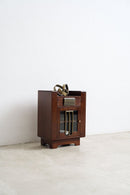 A converted Art Deco speaker cabinet  with a glass-front lower door and brass hardware, standing on a light concrete floor against a white wall.