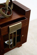A close-up of a dark brown wooden cabinet with brass hardware, including a ribbed pull handle and a decorative brass sculpture on top, set against a light beige background.