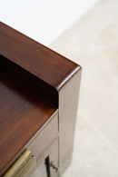 Close-up of a dark brown wooden furniture piece showing the corner of the tabletop and drawer front, with a brass pull handle and a light beige floor in the background.