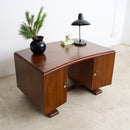 An Art Deco curved wooden desk with a warm brown finish, styled with a black lamp, a small green plant in a black pot, and a small box on a light beige floor against a white wall.