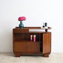 An Art Deco wooden sideboard cabinet with a warm brown finish, styled with a blue vase of pink flowers and small decorative items on top, set against a plain white wall and light concrete floor.