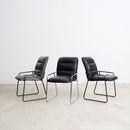 Three modern leather dining chairs arranged side by side against a plain white wall and light concrete floor. The left chair is green, the centre and right chairs are in black - upholstered in full grain aniline leather.