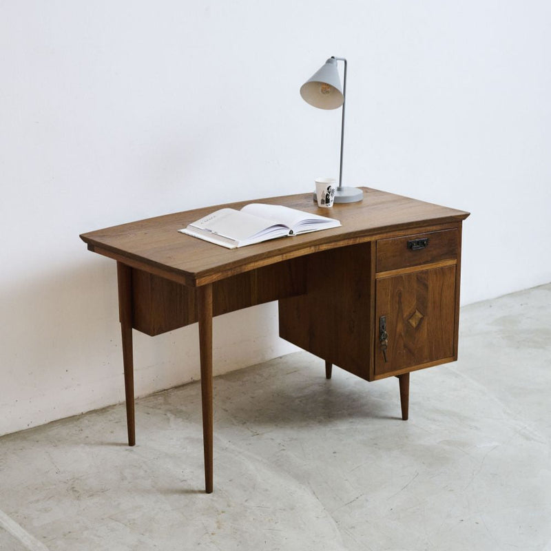Midcentury vintage teak wood desk with an open book and a lamp on a white floor and wall.